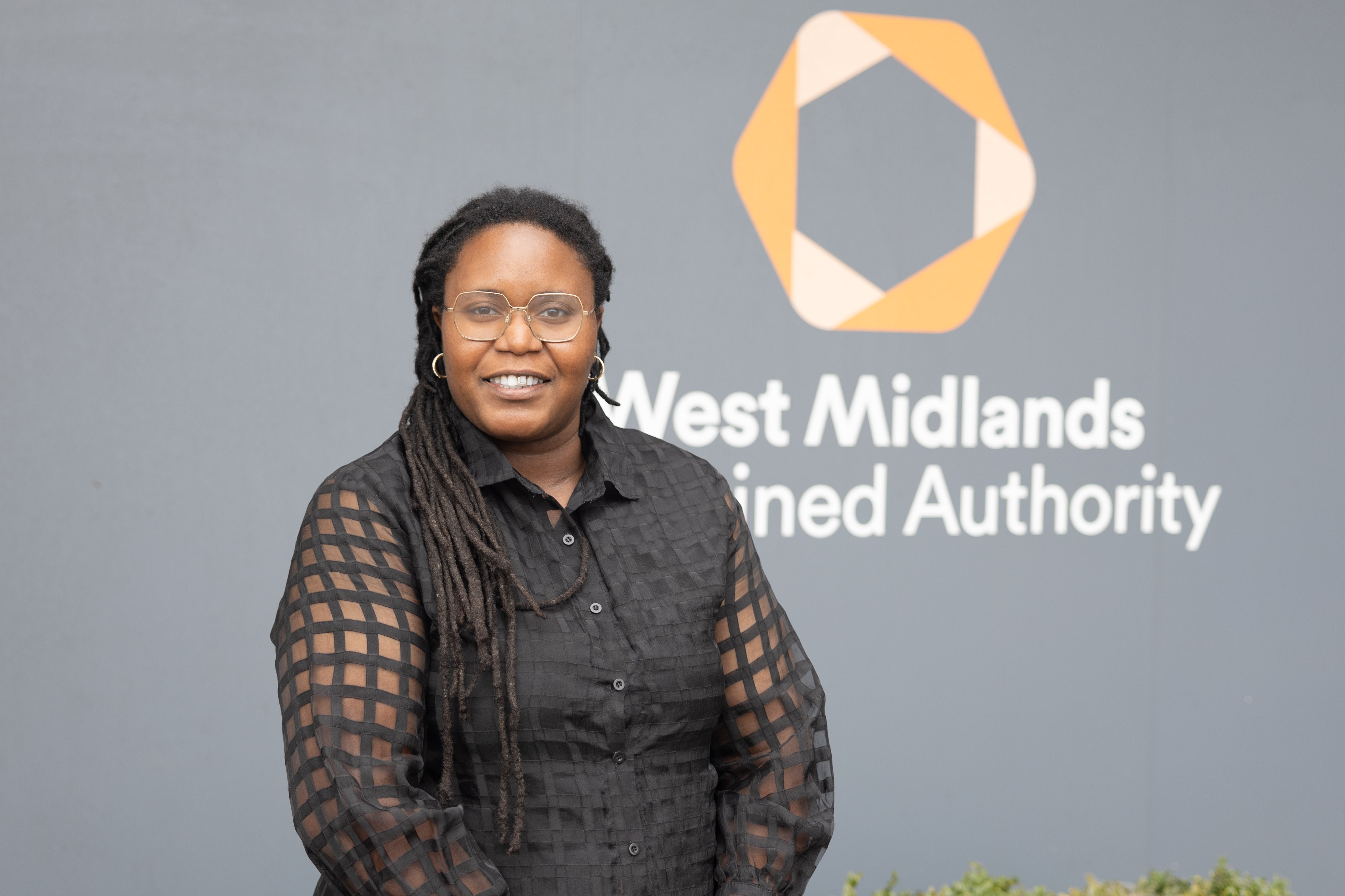 The image shows a woman standing outside an office. She is smiling and looking towards the camera. Behind her you can see a sign for West Midlands Combined Authority on the wall