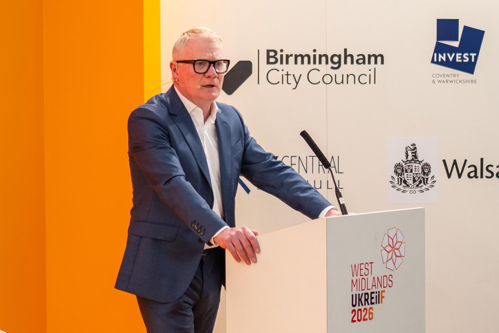 A man in a blue suit and glasses speaks at a podium with "West Midlands UKREiiF 2026" branding. Logos of Birmingham City Council and others are visible.
