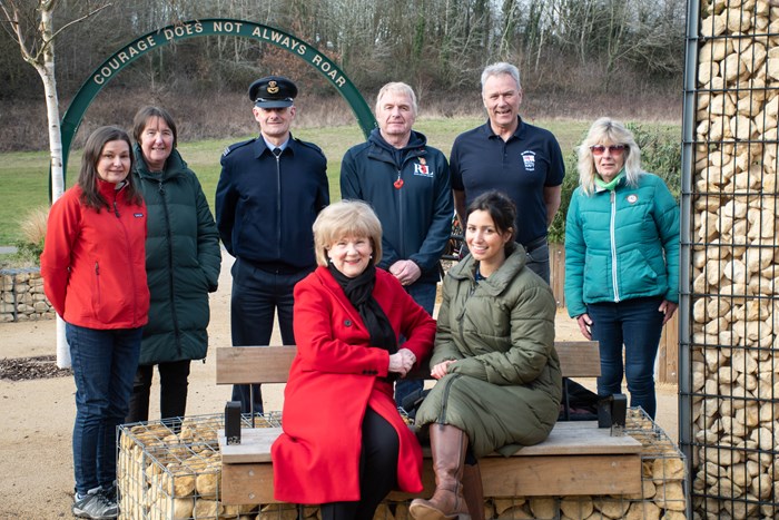 A group of 8 people stand or sit around a wooden bench in a garden with the entrance sign too the park displaying the words Courage Does Not Always Roar