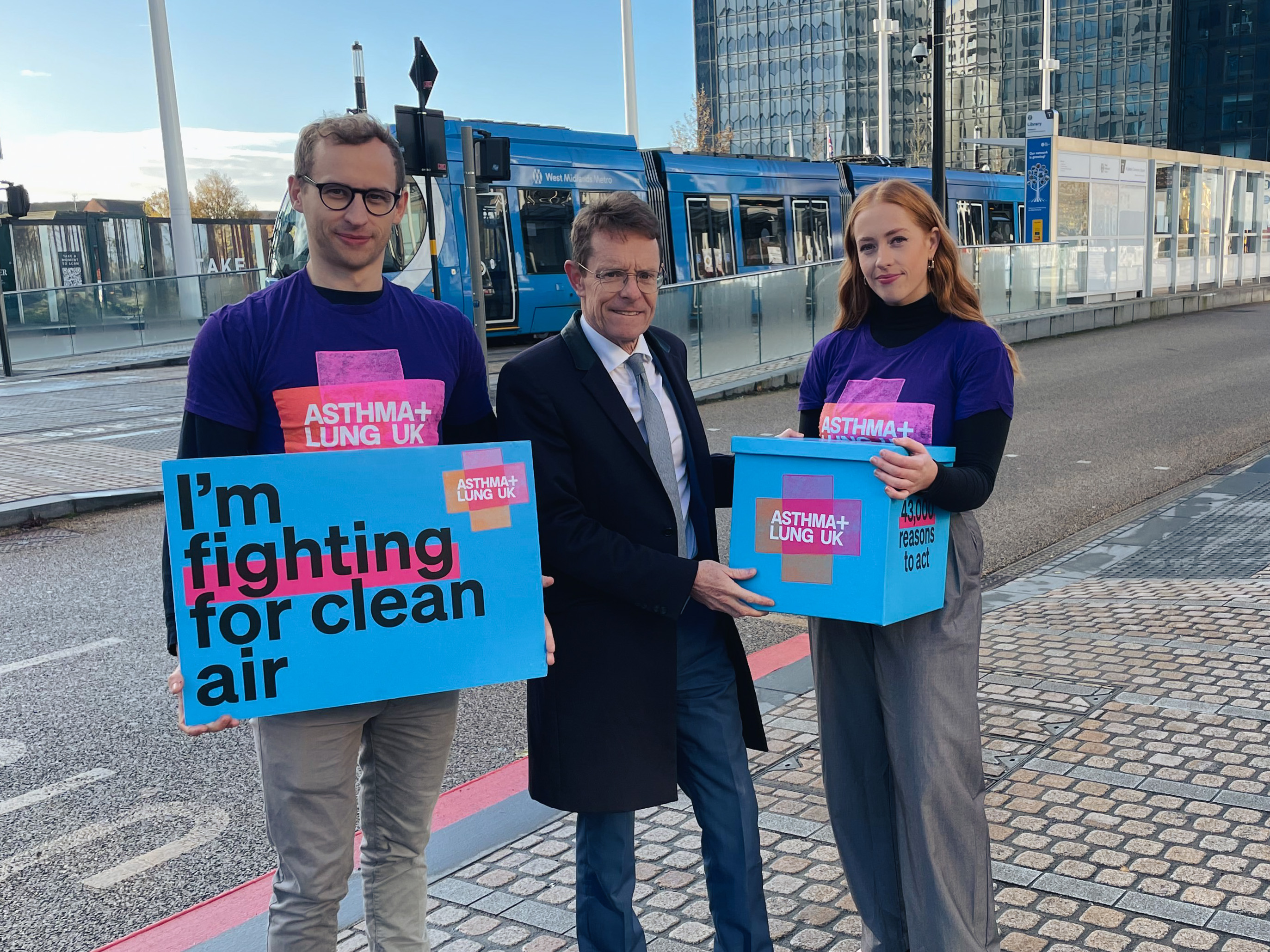 Three people looking at the camera as a petition is handed to the Mayor with a tram in the background.