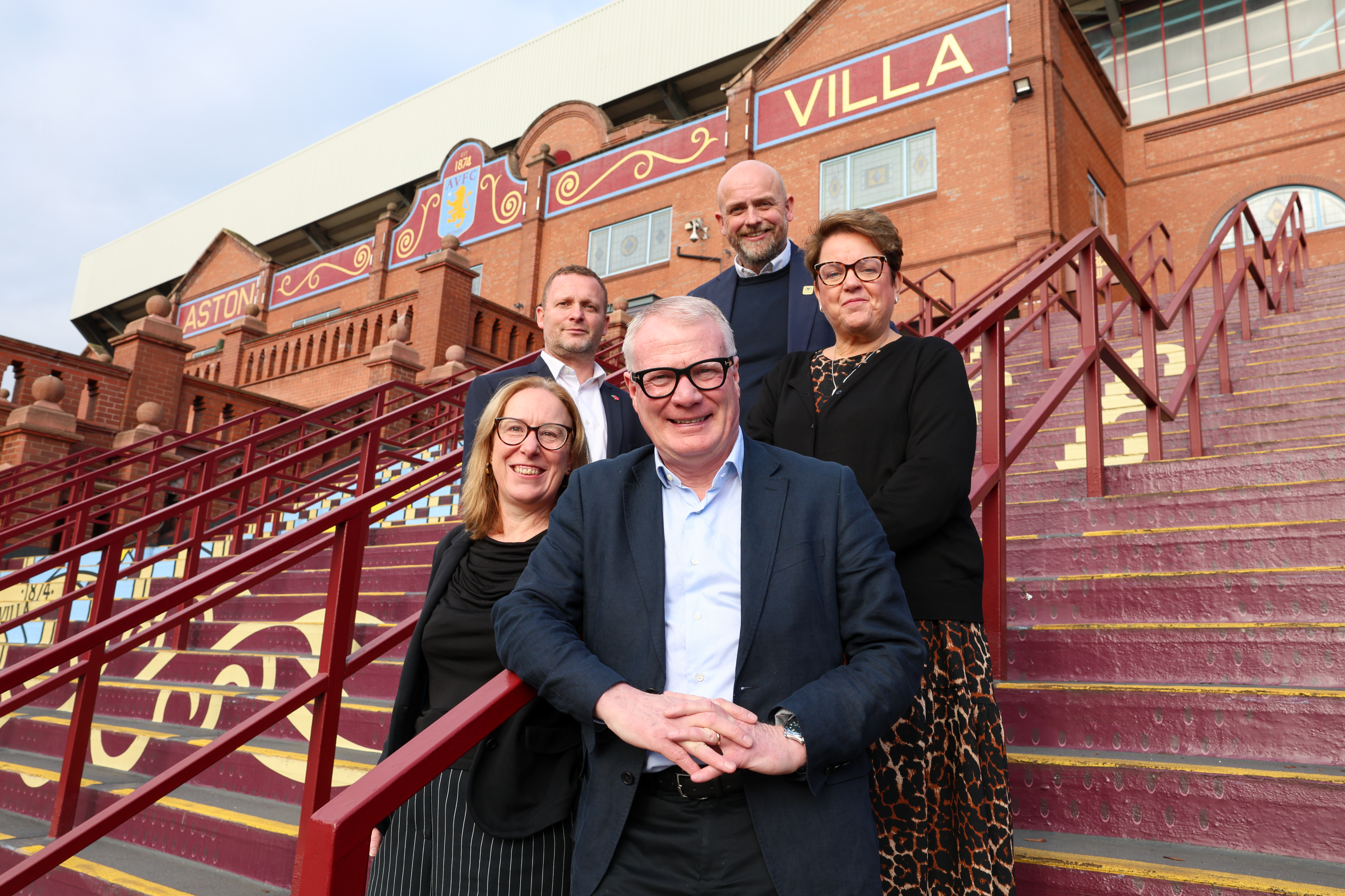 Five people, (left to right) Clare Sumner, (chief policy and impact officer, Football Association Premier League), Guy Rippon, (head of the Aston Villa Foundation), Mayor Richard Parker (centre, front), Will Clowes, (director of the Wolves Foundation), Helene Dearn, (executive director, Employment, Skills, Health and Communities, WMCA) standing on the steps outside Aston Villa Football club