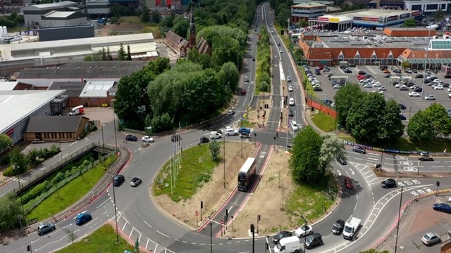 Aerial view looking down on a roundabout on a clear day. The roundabout has a road cutting through the middle linking two dual carriageways and a bus is driving through this. Other traffic circles around the outside of the roundabout.