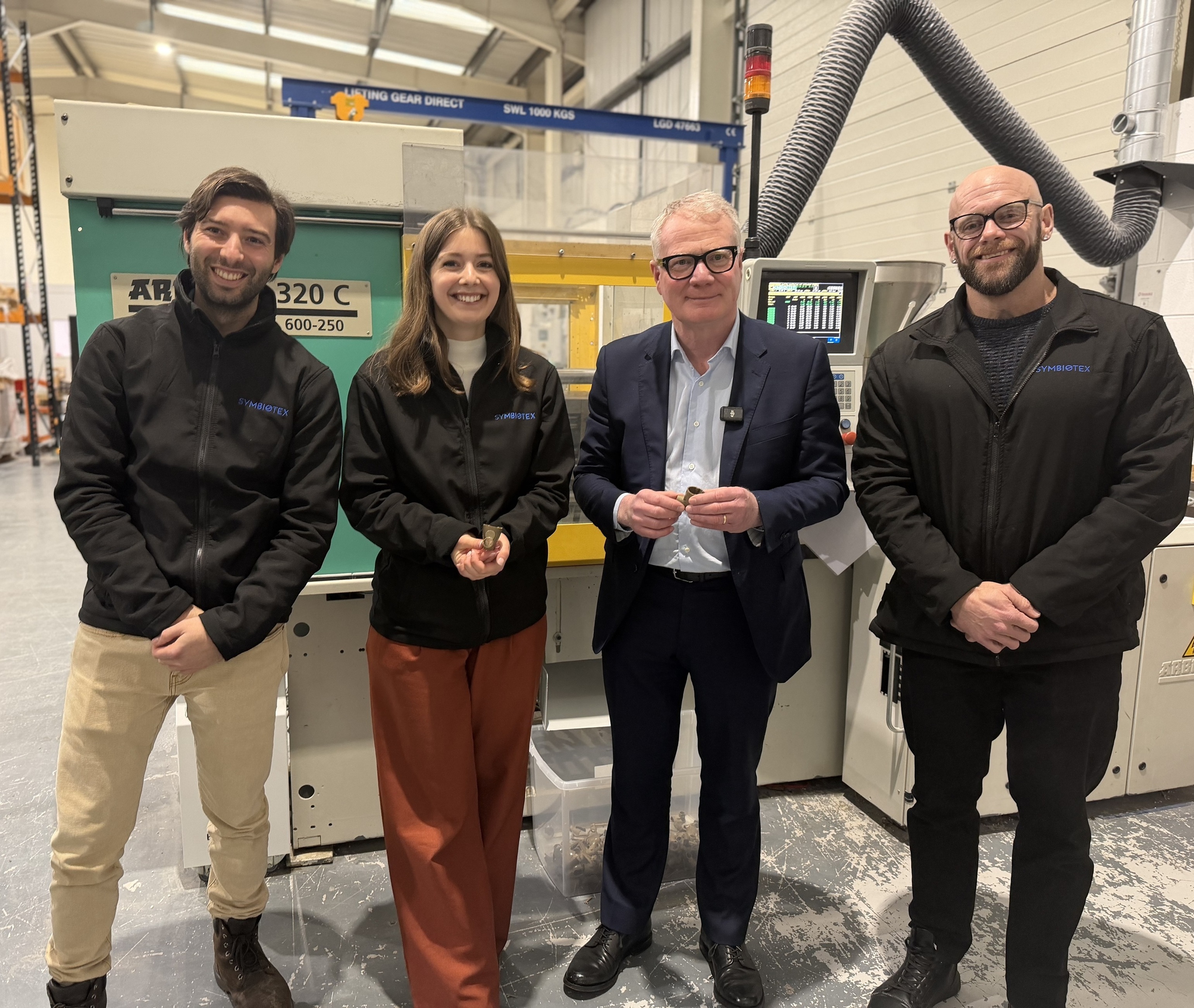 Four smiling colleagues pose beside industrial manufacturing equipment in a bright factory.