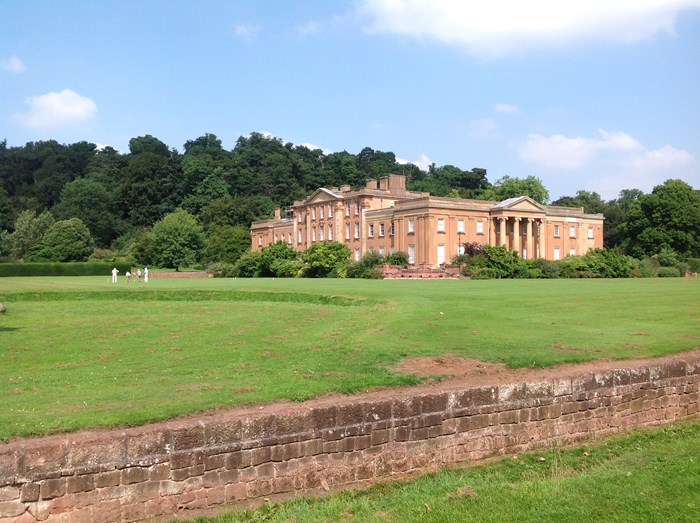 A grand country house to the right of the image with green parkland surrounding it and the remains of a moat in the foreground