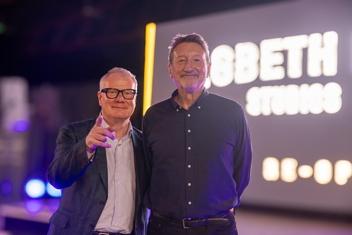 Two men standing together at an event, smiling at the camera. The man on the left wears glasses, a blazer, and a button-up shirt, and is pointing toward the camera. The man on the right is taller, with short hair and a beard, dressed in a dark button-up shirt. A brightly lit sign in the background reads “DIGBETH STUDIOS.”