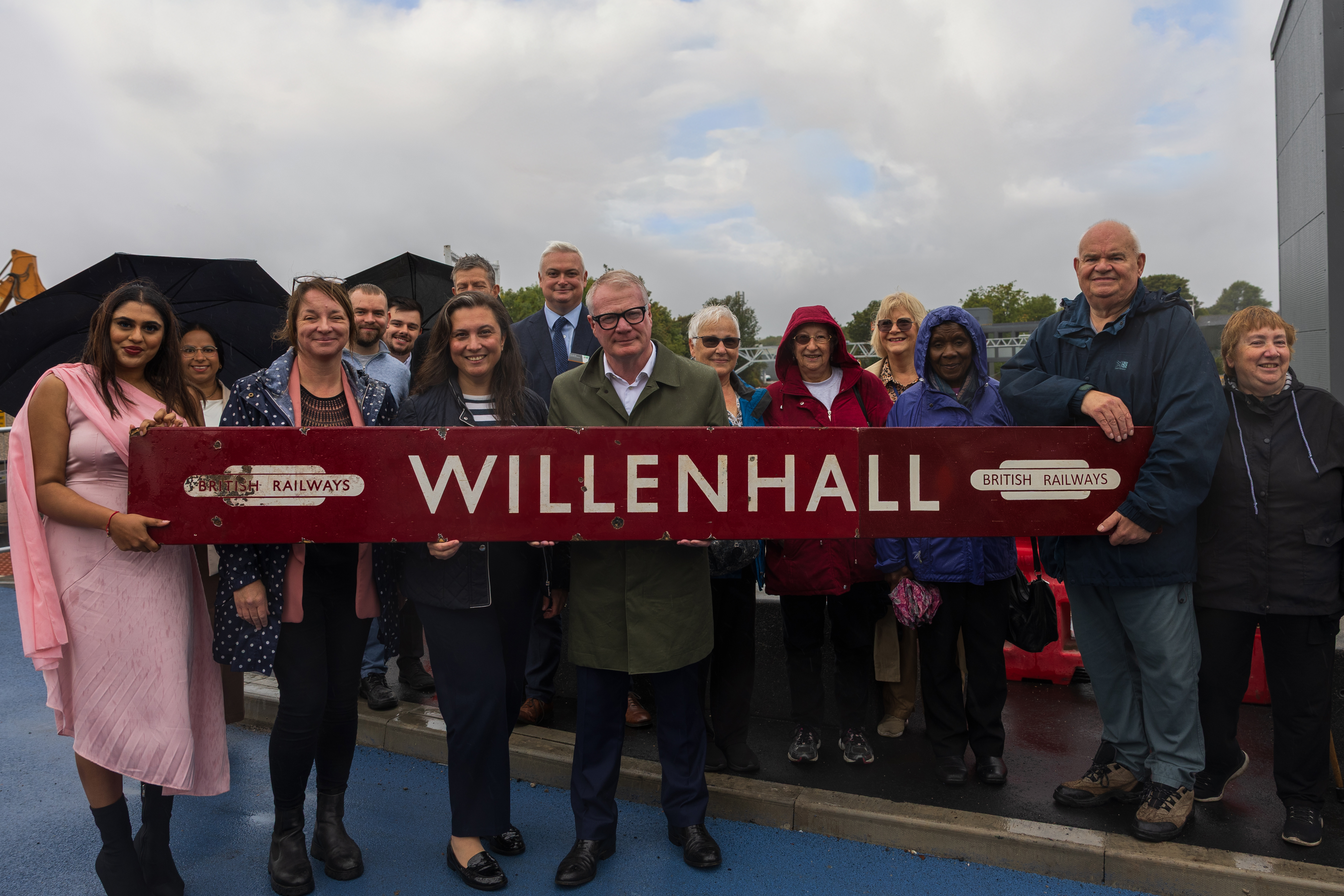 A group of about a dozen people - members of the Willenhall community - are gathered on a grey cloudy day. At the front of the group a large nine foot long sign saying "Willenhall" and British Railways is being held. The sign is a deep red with white writing. In the centre of the group is Mayor of the West Midlands Richard Parker.