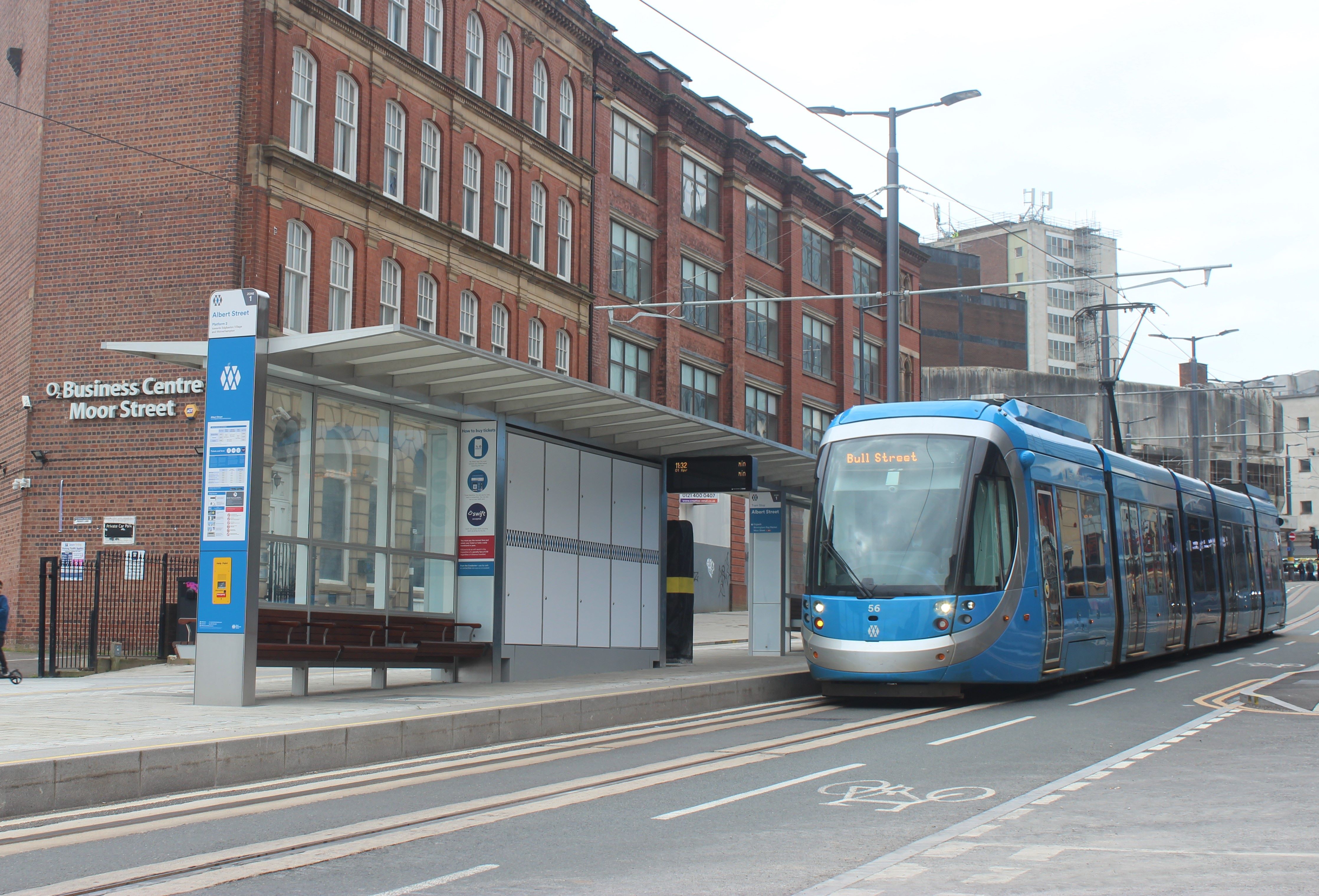 Blue tram approaches a tram stop platform in a busy city centre
