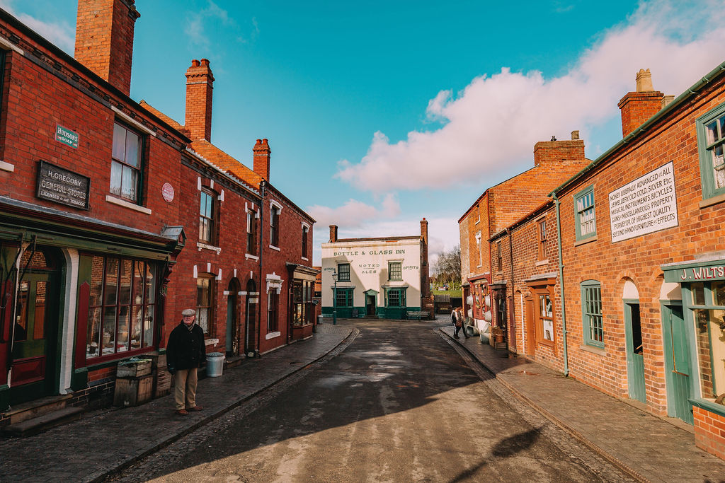 A landscape photograph featuring a High Street located at Dudley's Black Country Living Museum. In the scene, two actors are standing outside their respective shops.