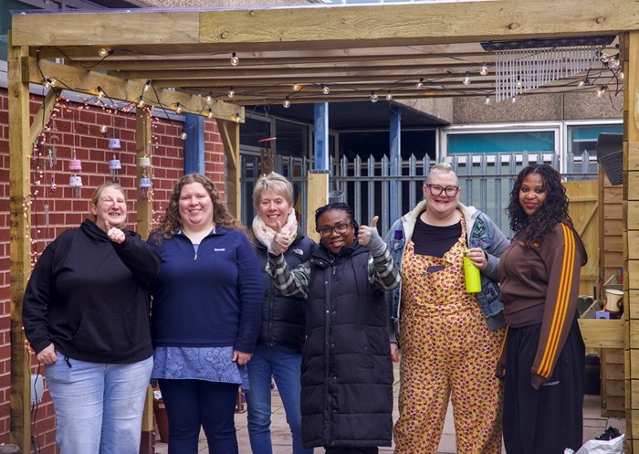 Six diverse women smile and pose beneath a wooden outdoor shelter, wearing colorful casual clothing