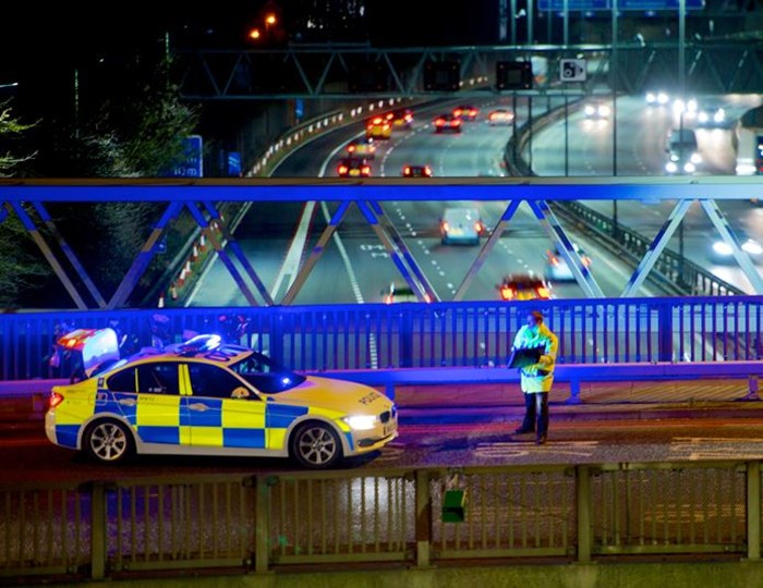 Police officer in yellow high-visibility jacket standing on a motorway bridge while monitoring traffic speeds below