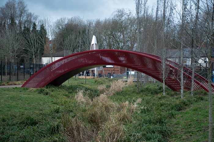 A bridge made from steel stretches over a waterway currently dried up with grass banks on either side and trees and blue sky in the background