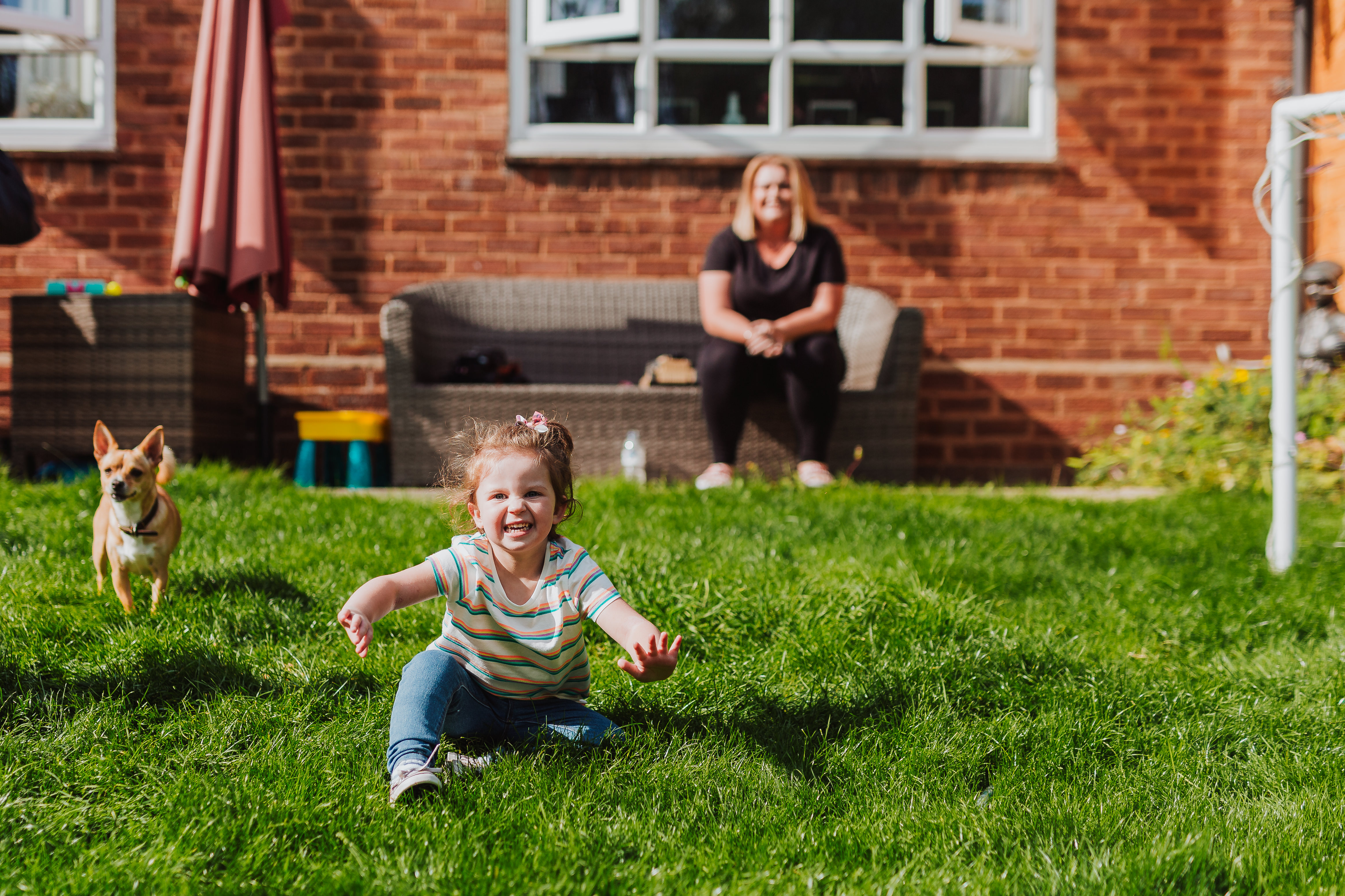 A woman sits on a bench in her garden in the distance of the image with a little girl, around 3 years old, plays on the lawn in front next to her dog