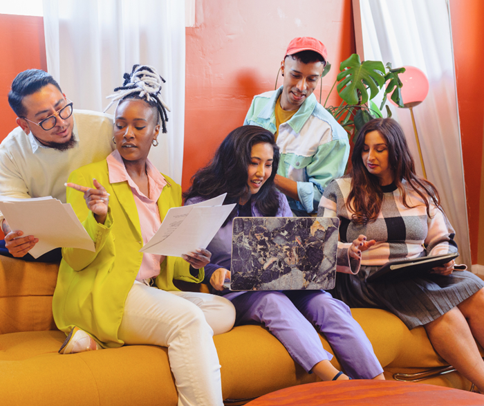 A group of 5 people sat on a bright yellow sofa, in front of bright orange walls. They have laptops and notebooks and are happily chatting.