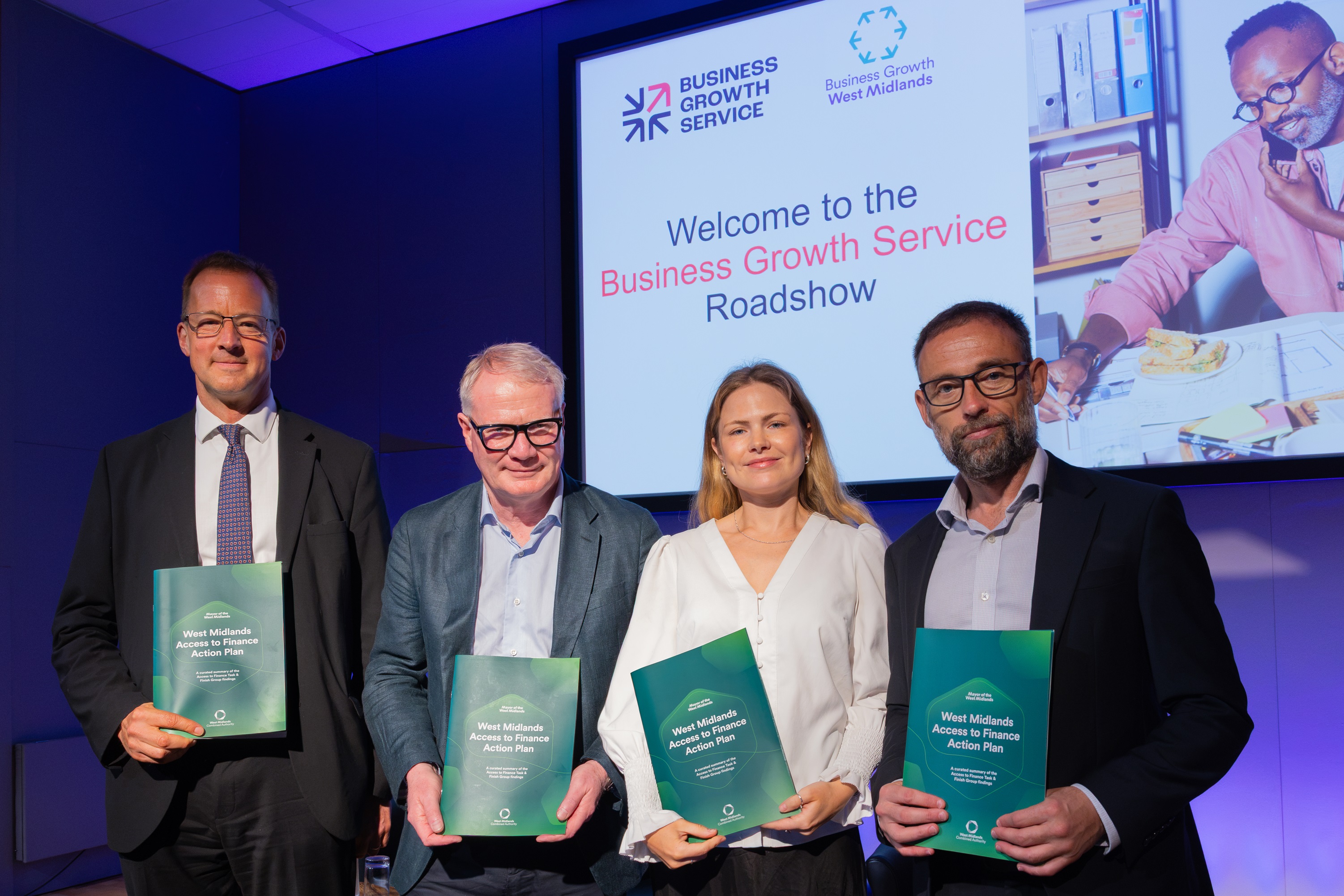 Four professional individuals holding booklets while standing in front of a screen that reads "Welcome to the Business Growth Service Roadshow."