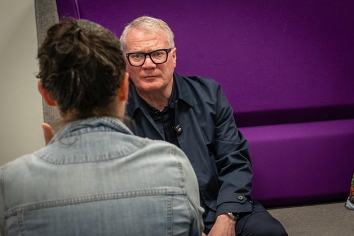 A man in glasses and a dark jacket speaks with a person in a denim jacket, seated against a purple wall in a focused conversation.
