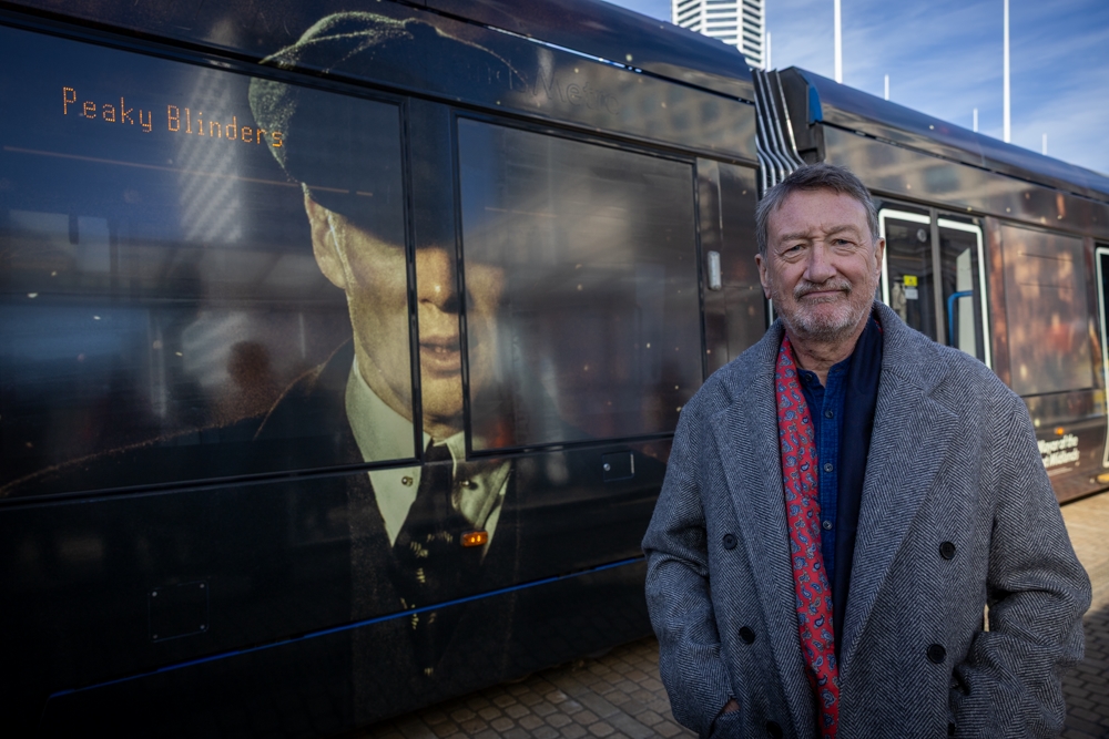 Steven Knight standing with, over his should, the side of a tram bearing a large image of Tommy Shelby, the lead character from Peaky Blinders, wearing his distinctive cap