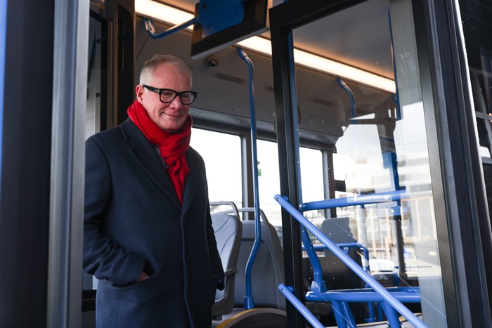 A man, Mayor Richard Parker, is standing in the doorway of a blue trimmed bus about to get off. He's wearing a black over coat and red scarf