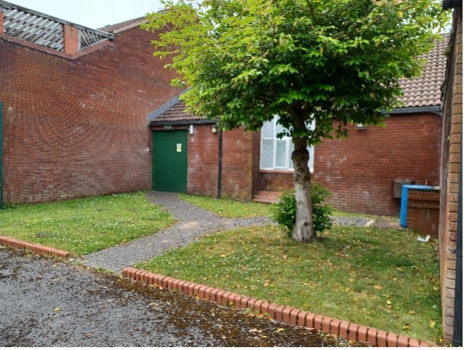 A small grassed area outside a brick built building with a tree to the right