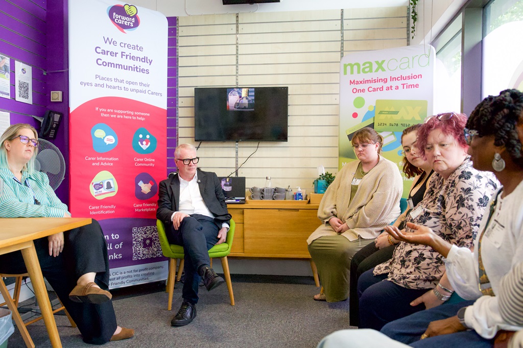 A diverse group of adults sits in a community meeting, listening and talking in a welcoming room with “Carer Friendly Communities” and “Max Card” posters.
