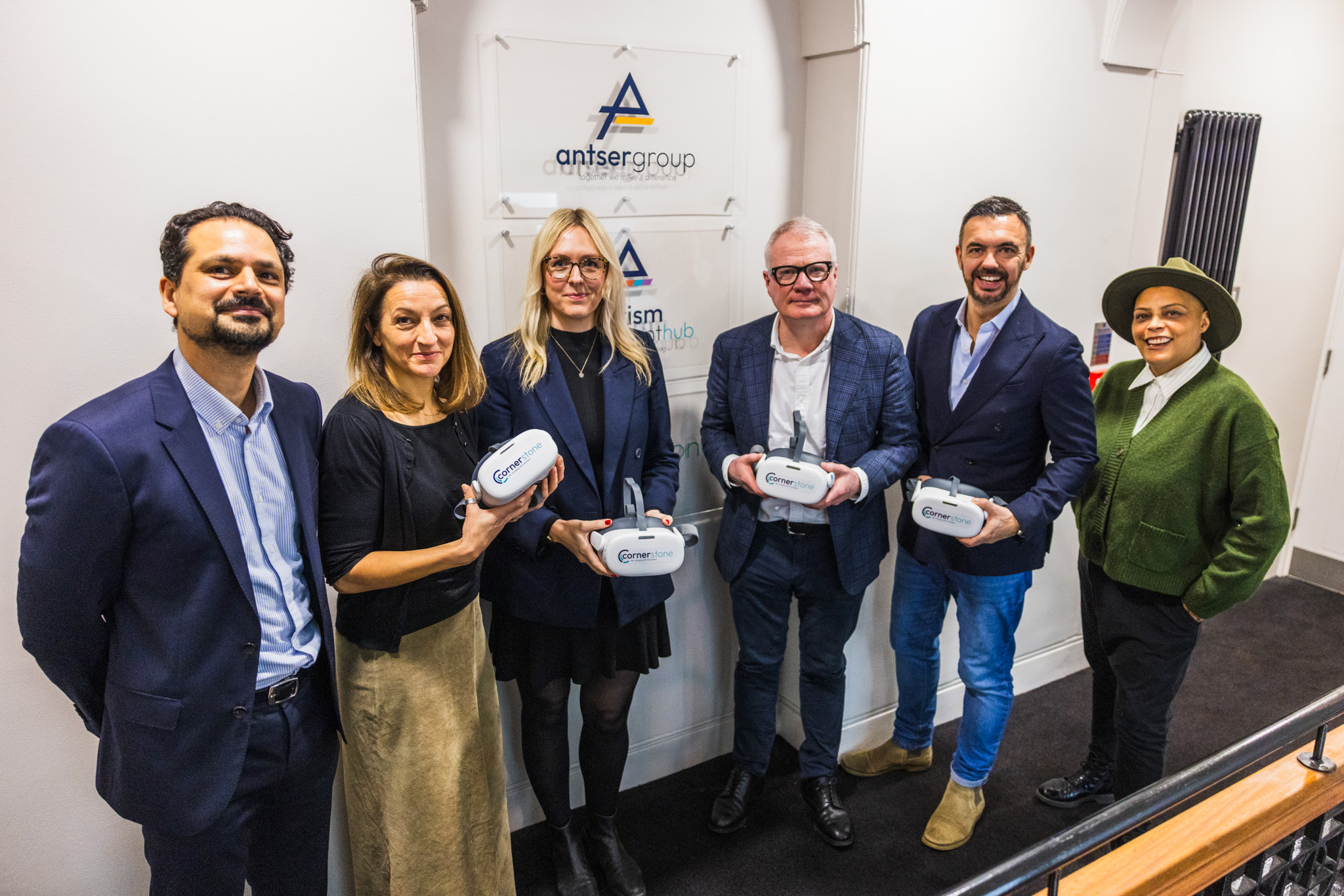 A diverse group of six people stands indoors, smiling and holding virtual reality headsets. They are in front of signs reading "Antser Group" on a white wall.