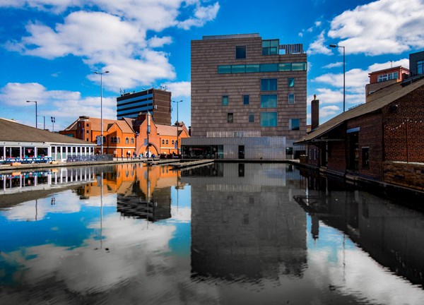 Canal with reflections of modern and red-brick buildings, waterside restaurants, and blue sky with clouds.
