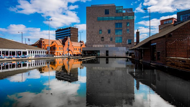 Canal with reflections of modern and red-brick buildings, waterside restaurants, and blue sky with clouds.