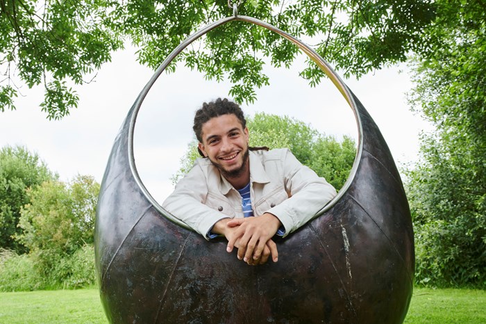 A man lending with his arms in the middle of a sculpture which is based within a meadow-style field surrounded by trees