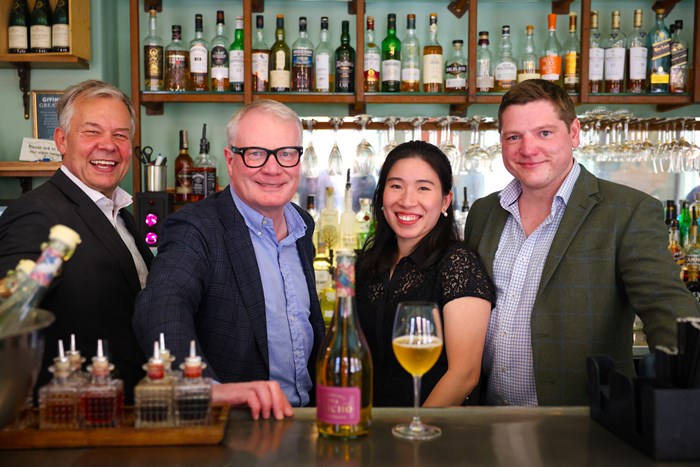 Line up of four people behind a bar - with shelves of bottles on the back wall. The Bottle of Siacho and a full wine with the light coloured drink sit on the bar in front of them.glass