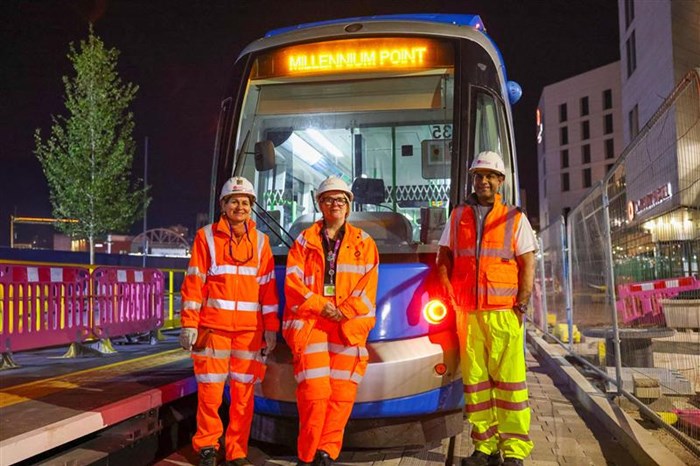 At night, three people in construction suits standing in front of a blue tram with Millennium Point on the destination board