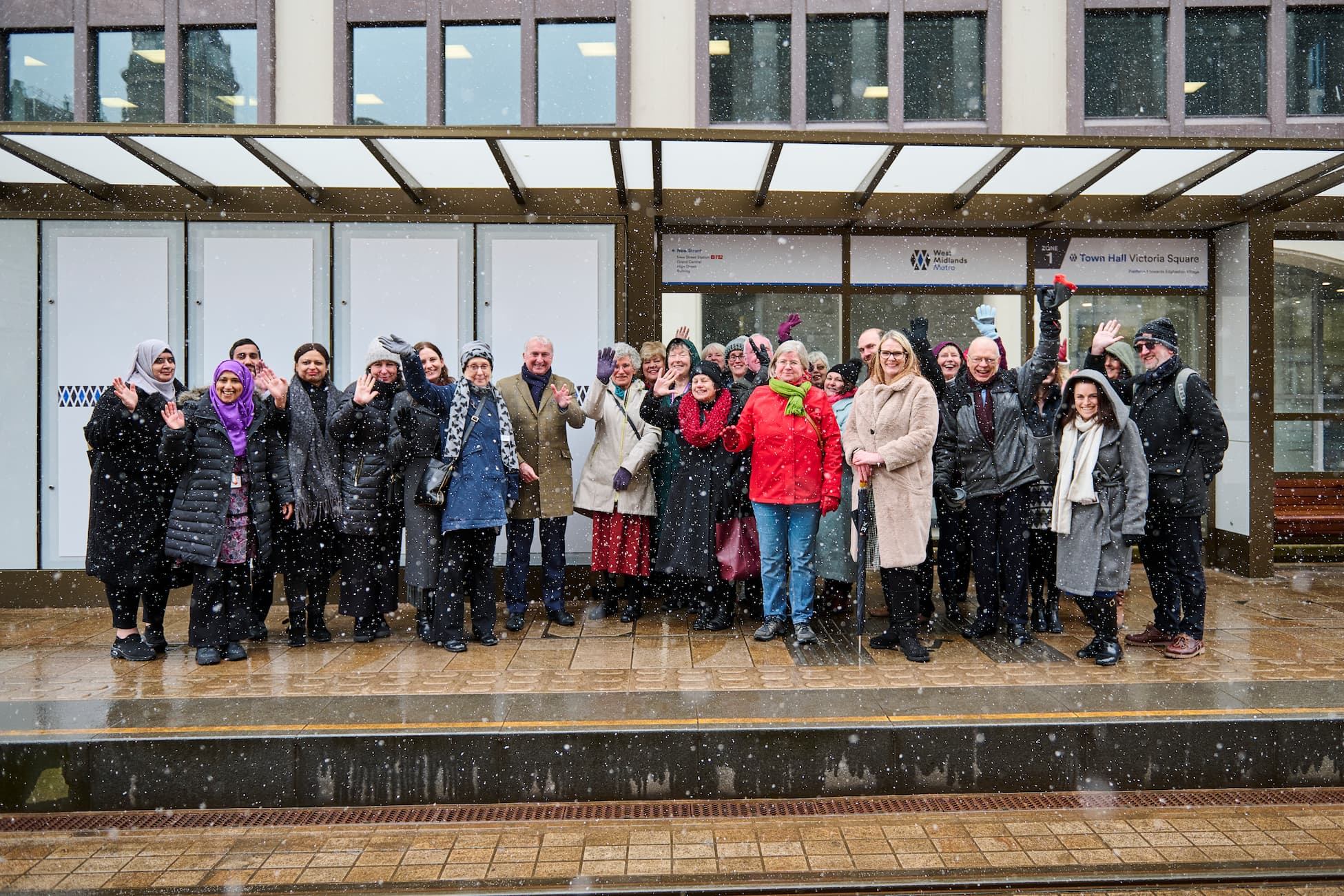 A group of people standing outside a building in snowy weather, smiling and waving, with a glass canopy overhead and signage on the wall behind them