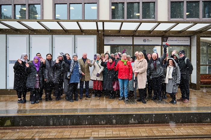 A group of people standing outside a building in snowy weather, smiling and waving, with a glass canopy overhead and signage on the wall behind them