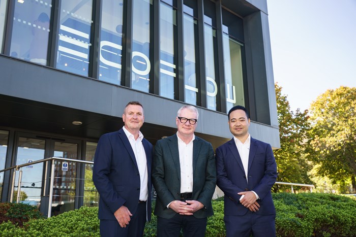 Three men in business attire standing outside a modern office building with large windows, greenery in the foreground, and trees in the background on a sunny day