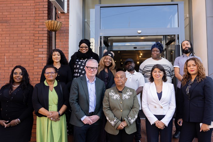 A group of thirteen people pose for a photo outside a building with a glass entrance. They are dressed in business and casual attire, standing and sitting in two rows.