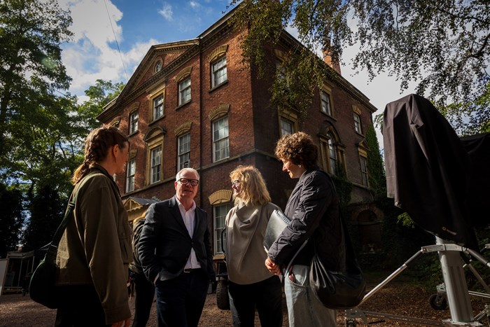 Three people talk beneath a towering ivy-covered red-brick building, framed by trees and studio equipment; warm sunlight gives the scene a candid, thoughtful mood.