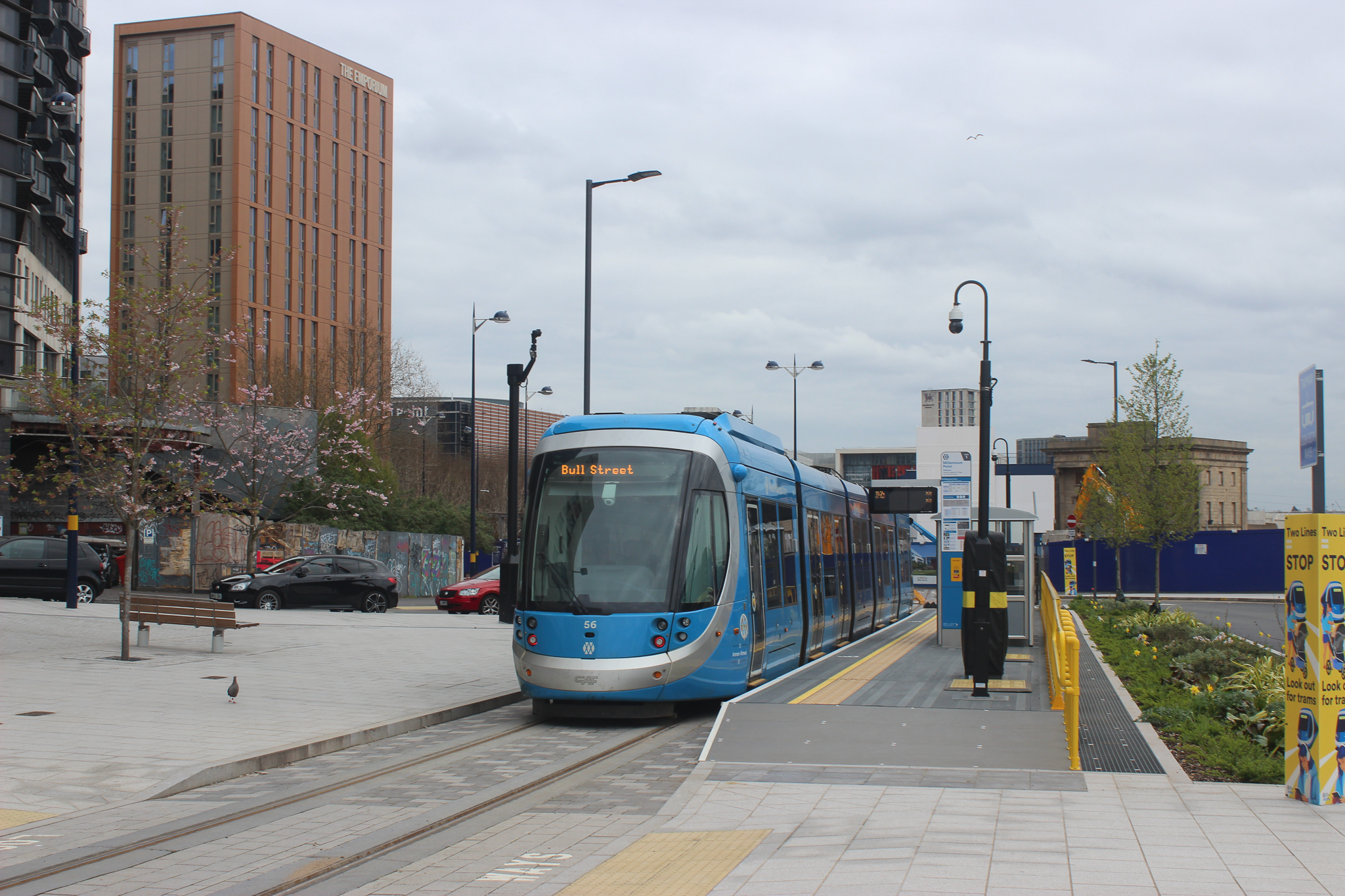 Blue tram parked at a tram stop platform with a tower block and city buildings behind