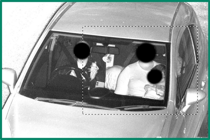 Black and white security camera image shot through car front window shows passenger with small child on lap and no seatbelt on