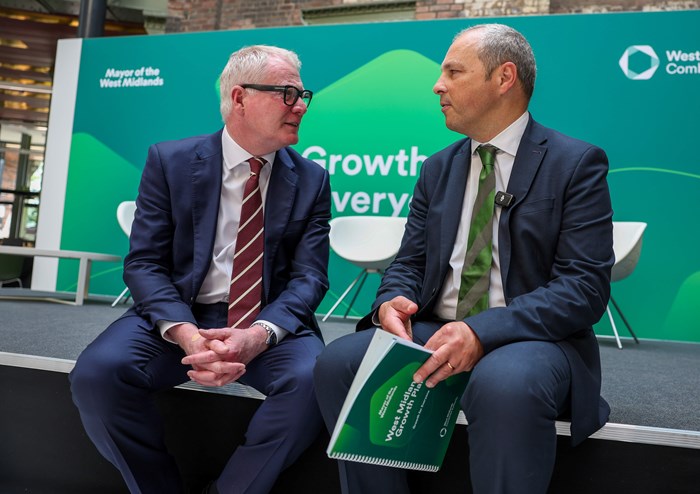 Two men sit on a stage at a conference centre. Behind them is a board that says Growth for Everyone with the logos for Mayor of the West Midlands and West Midlands Combined Authority