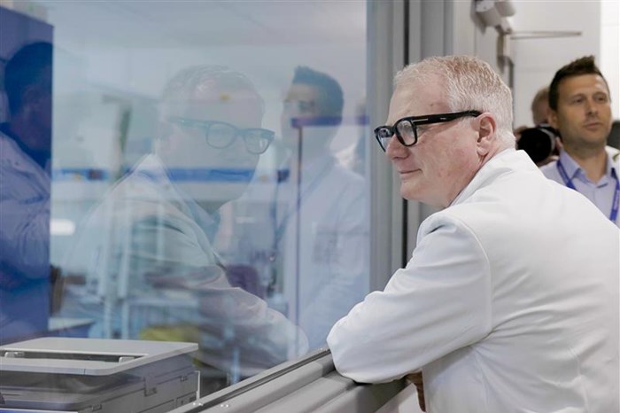 The Mayor is on the right of the picture, he is wearing a white lab coat and, seen from the side, he is leaning on a window sill and looking left. He is viewing a laboratory through a window.