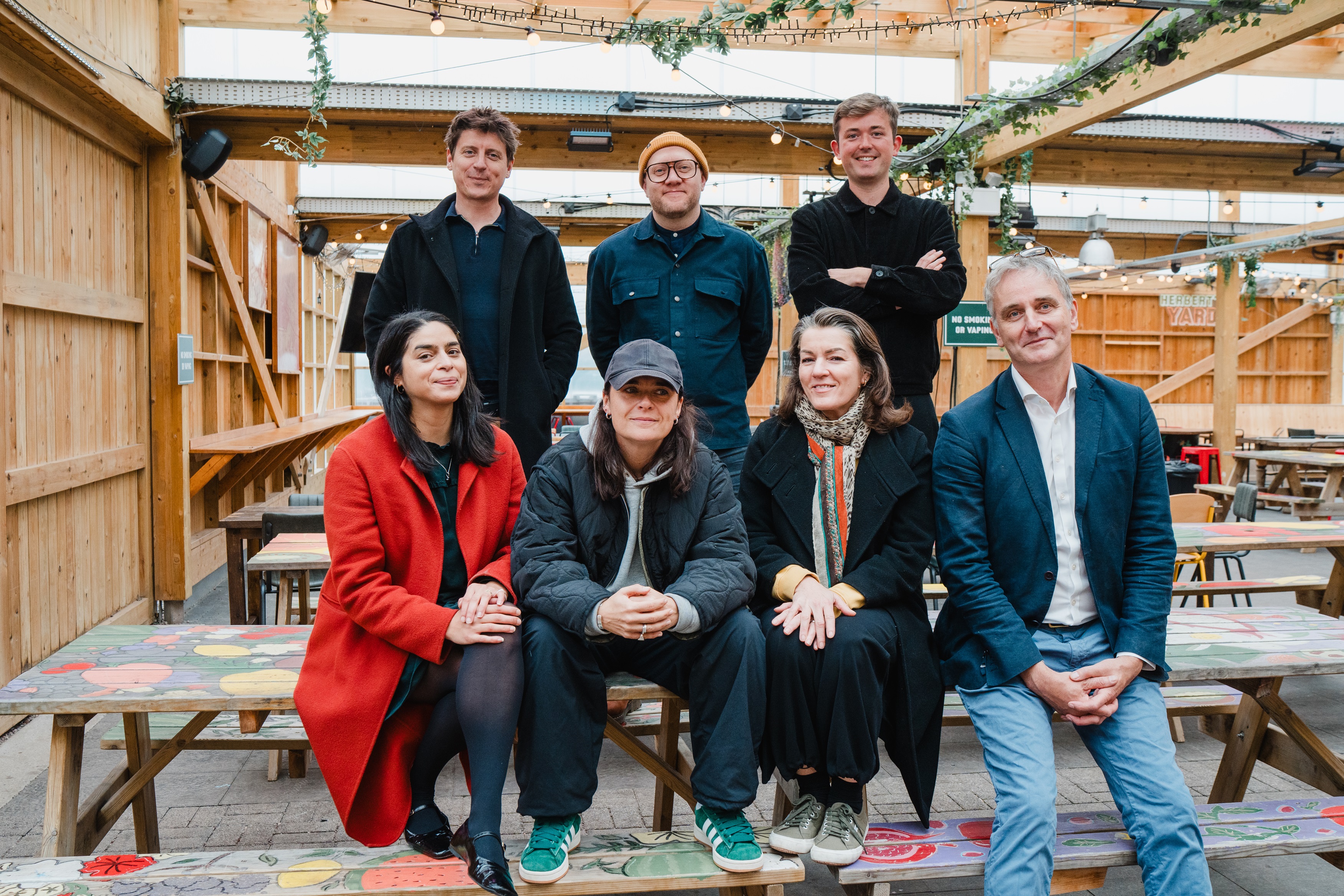 A group of seven people smile at the camera indoors, seated and standing at colorful picnic tables, with a casual and friendly atmosphere.