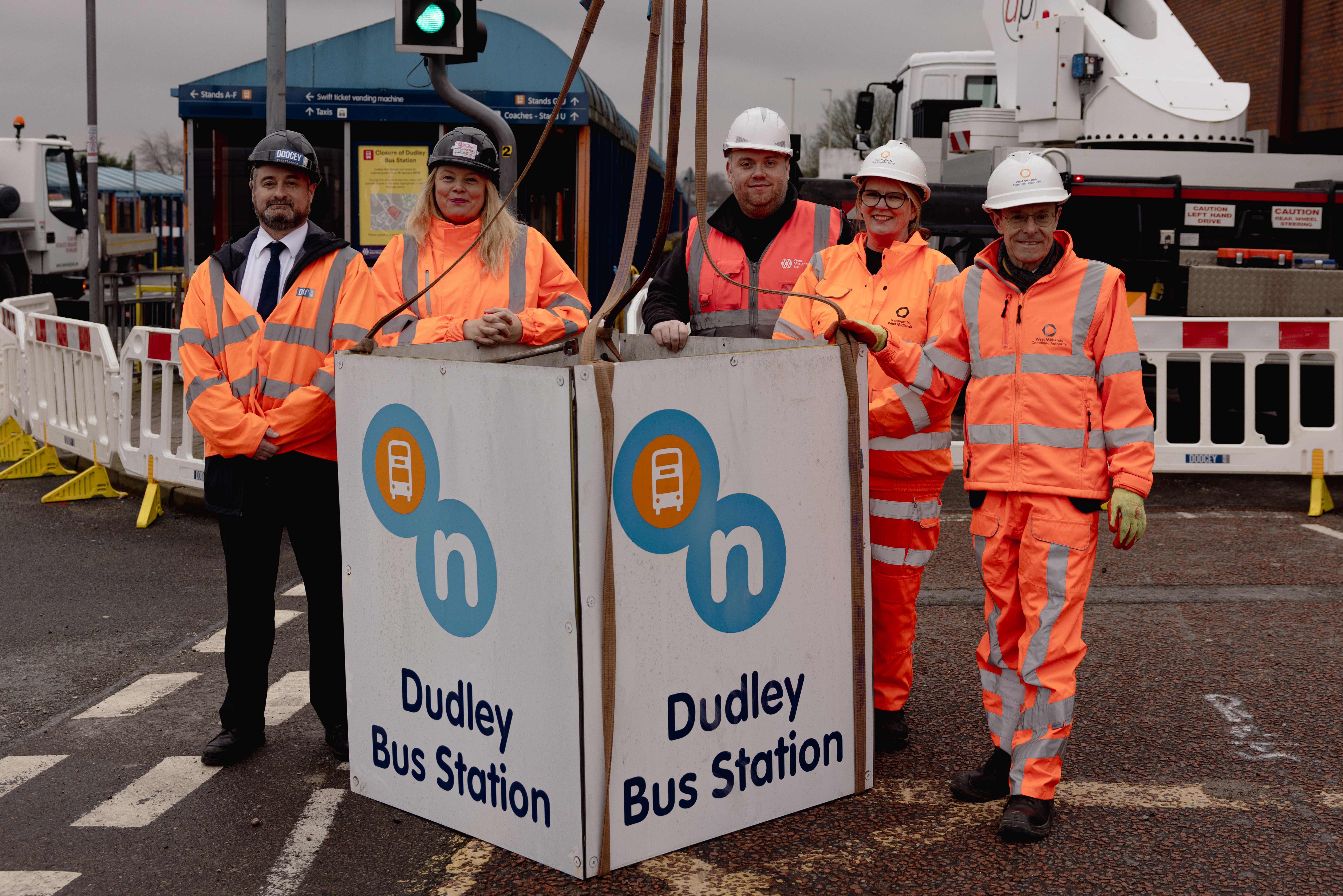 Five people in bright orange hi-vis construction outfits with the old Bus Station sign taken from a pole high above the bus station