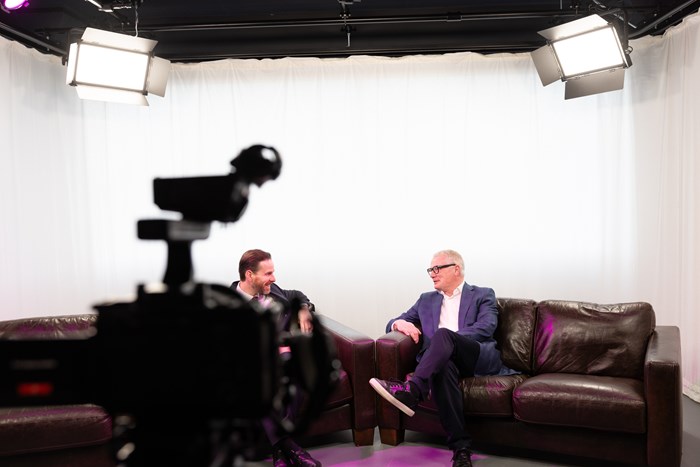 Two people sitting on a sofa in a studio environment with a camera in the foreground filming them.