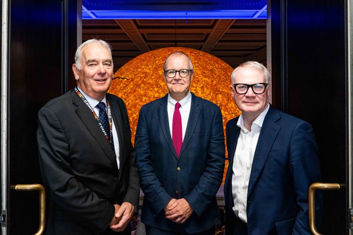 Three men in a formal pose and environment - the man on the left wears a black suit and a tie with a colorful pattern. The man in the middle wears a blue suit and glasses, and the man on the right wears glasses and a white shirt with a dark jacket.