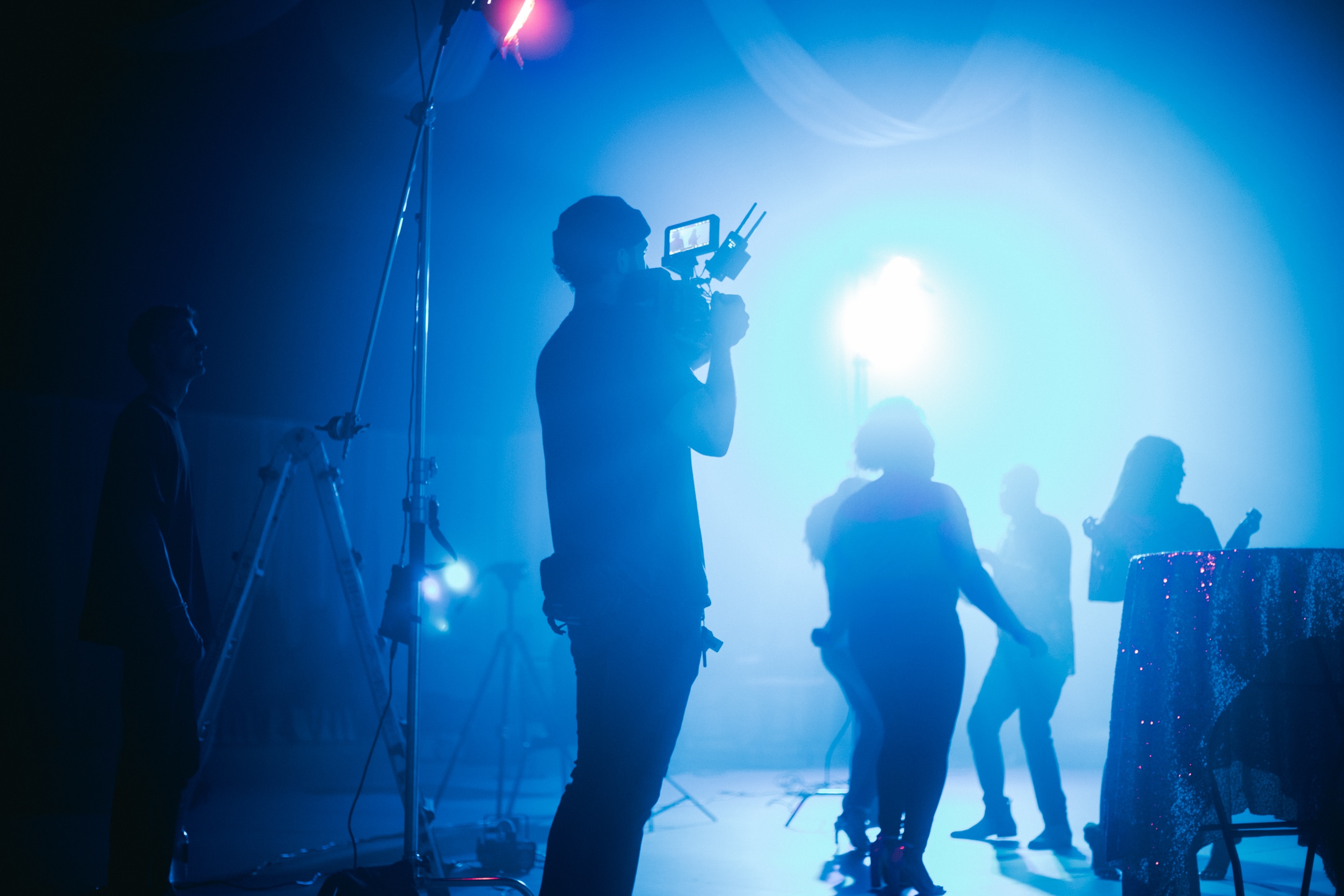 A photograph capturing a behind-the-scenes film shoot. The blue lighting in the room cuts through the dark, foggy atmosphere of unknown actors dancing in the scene.