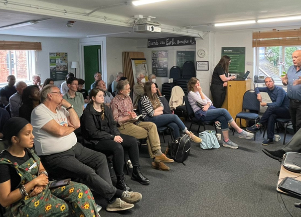 Residents seated in a community meeting room listening to a speaker at the West Midlands Greener Together Forum, with a mix of attendees facing the front while a presenter speaks and another person stands at a lectern.