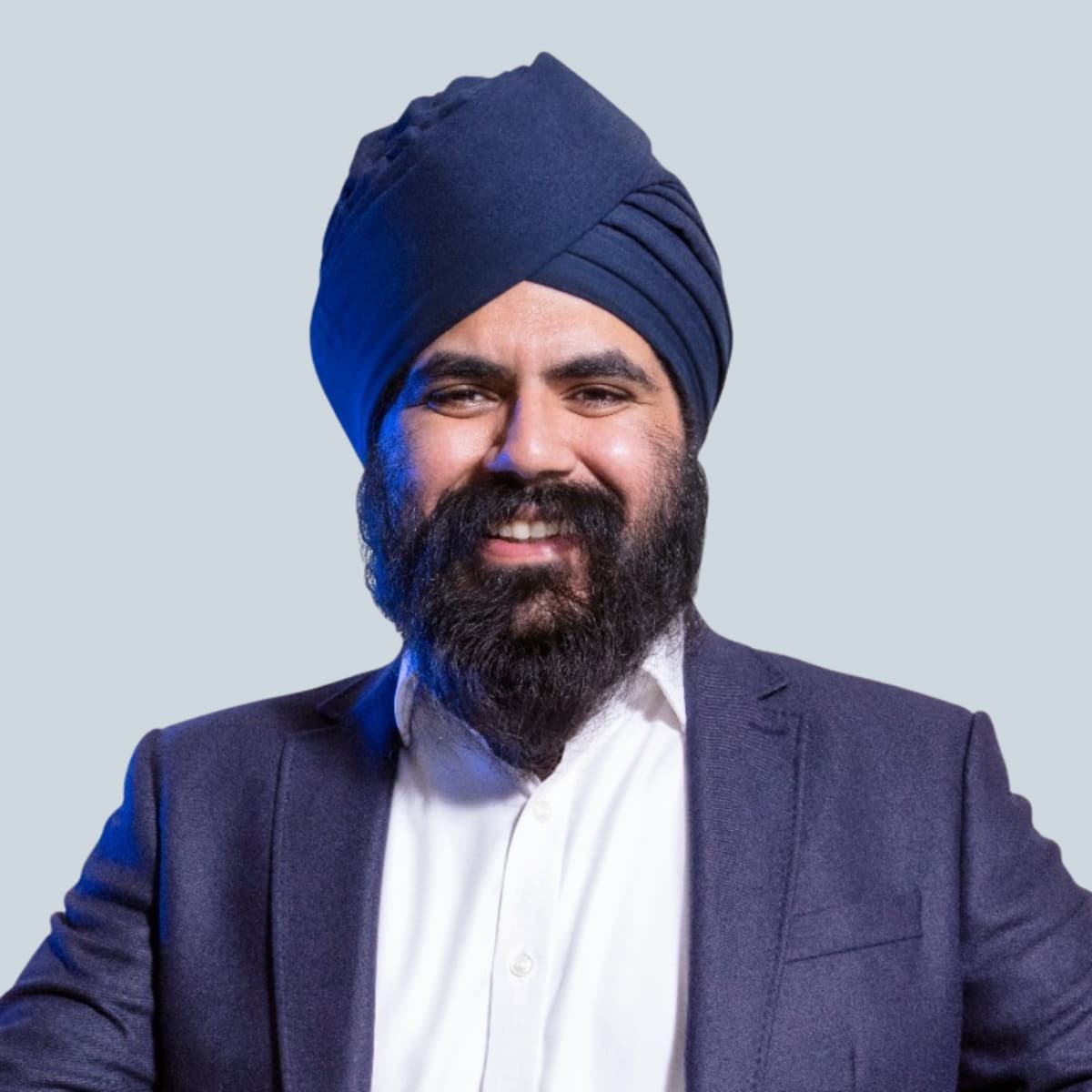 Smiling man with a beard and navy turban, wearing a white shirt and dark blazer, standing against a light gray background. Confident and approachable.