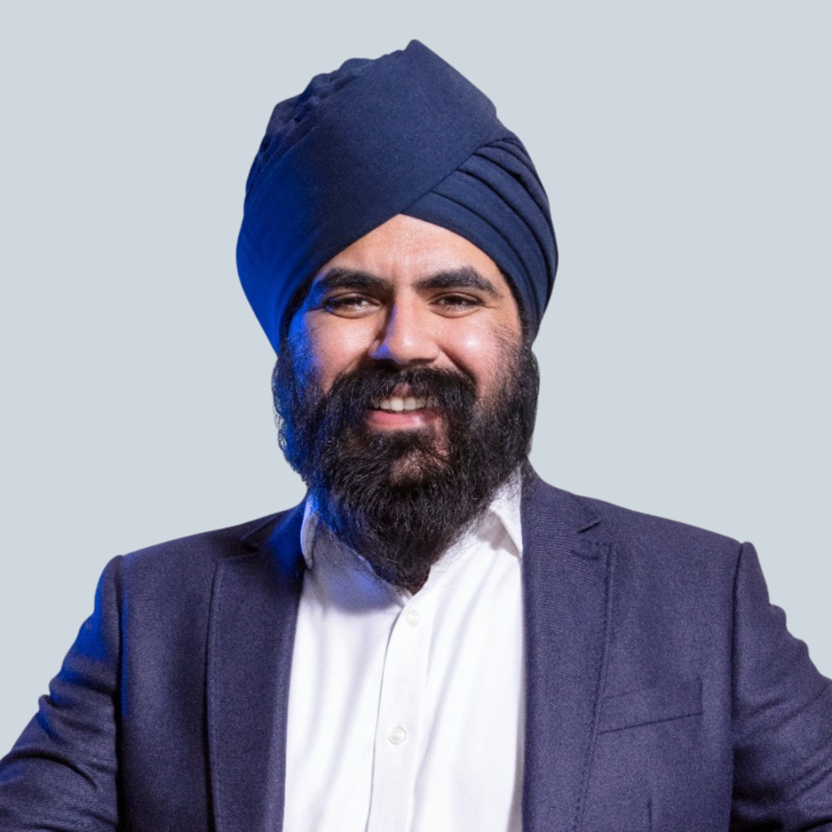 Smiling man with a beard and navy turban, wearing a white shirt and dark blazer, standing against a light gray background. Confident and approachable.