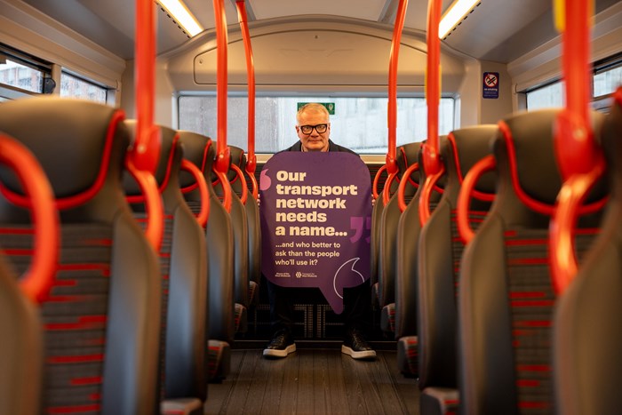 Mayor sits on the back seat of a bus with a sign saying our transport network needs a name