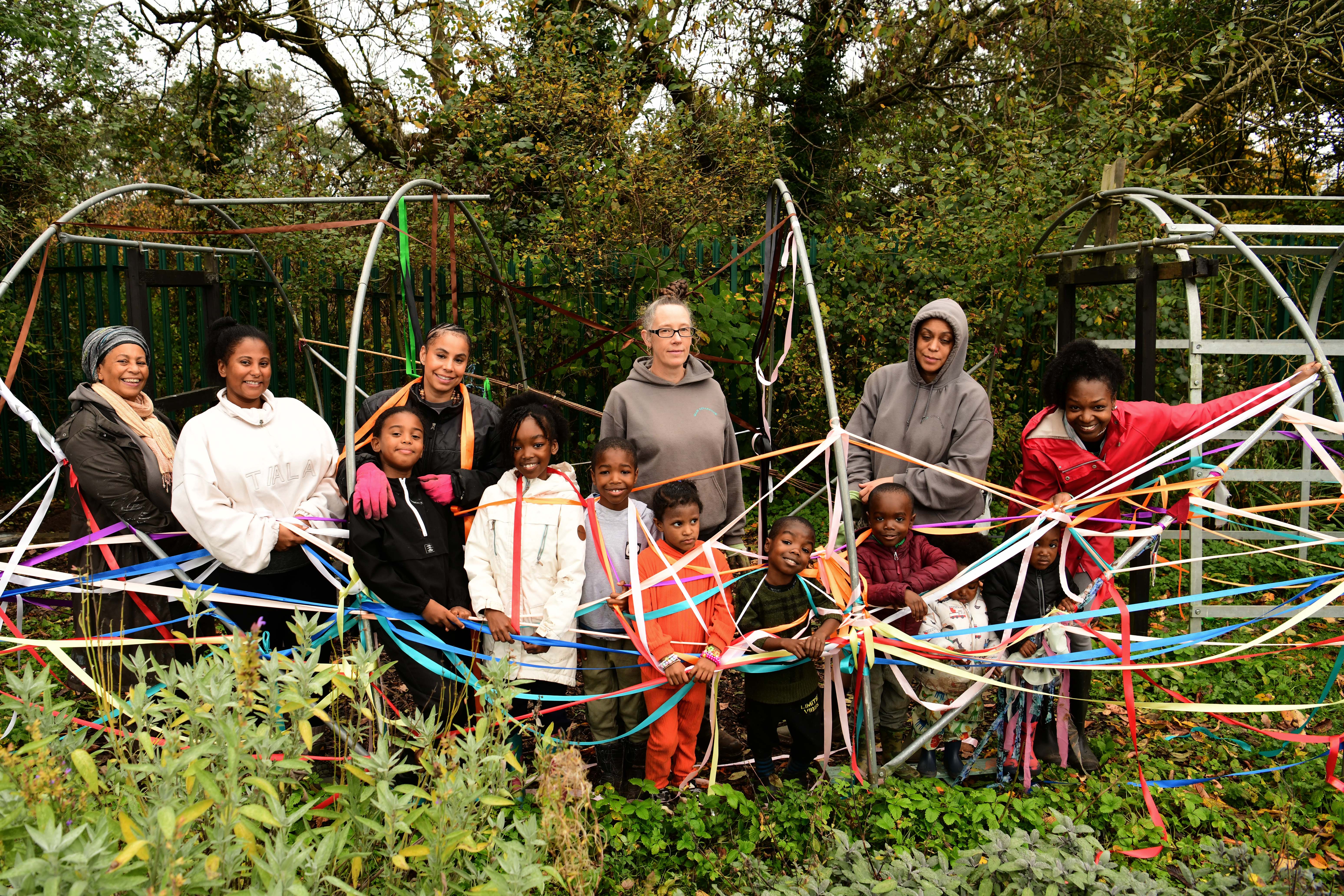 A group portrait of the Glue Collective posing with families and children outdoors in a green area reminiscent of an allotment.