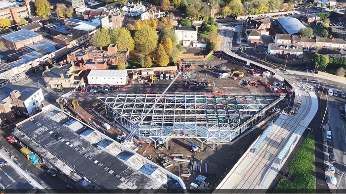 Aerial shot overlooking a large town centre construction site. A trapezium shaped steel frame dominates the image. And tram line runs along the side with a stop next to the frame building.