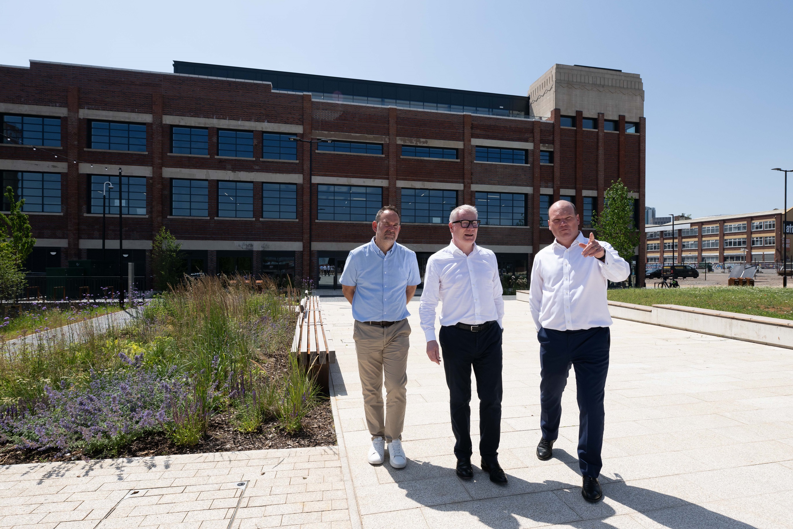 Three men walk on a sunny pathway beside a modern brick building, surrounded by greenery.